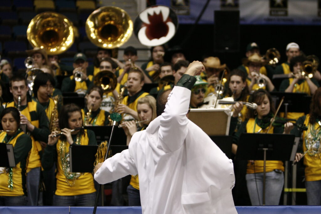 Dr. Michael Nickens directs the 2007 Green Machine Pep Band in a white suit. His right hand is raised in the air to direct a musical accent. The band is in gold basketball jerseys and is playing a song.