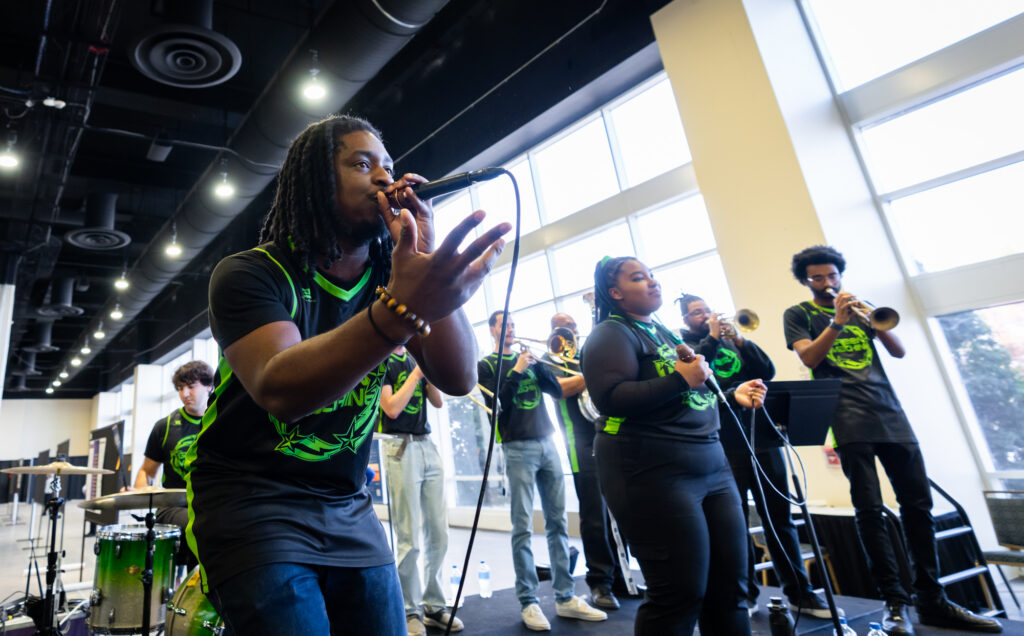 Members of Green and Gold Soul perform in a long industrial style office space. They are wearing black jerseys with green print.