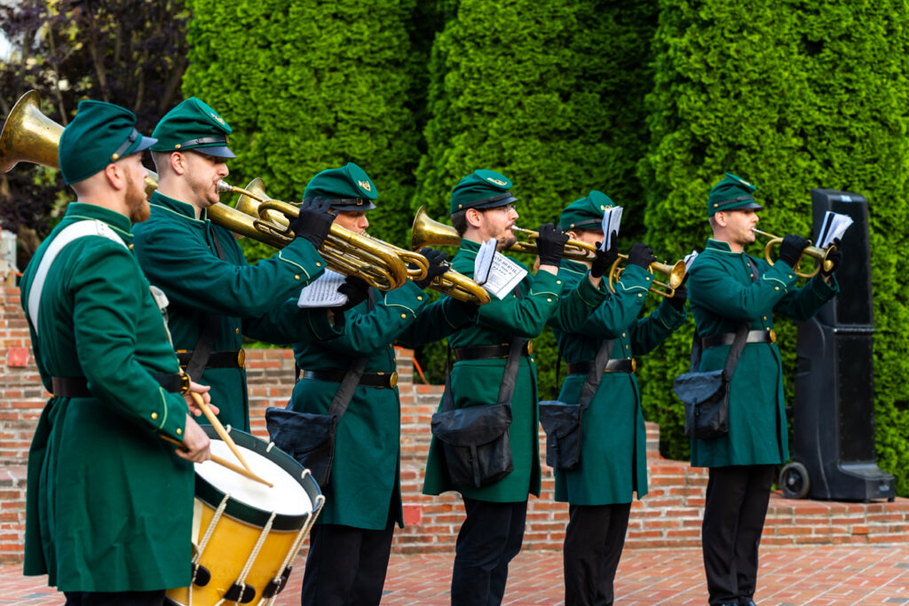 In the blurred background, three very tall pine shrubs are well-trimmed and uniform. Six members of the 8th Regiment Band are standing in a row on a brick stage set in an outdoor auditorium. They are wearing green 19th century military band uniforms with green military covers, and black wool pants. The member closest to the photographer is playing snare drum. The rest of the members are playing 19th century brass instruments.