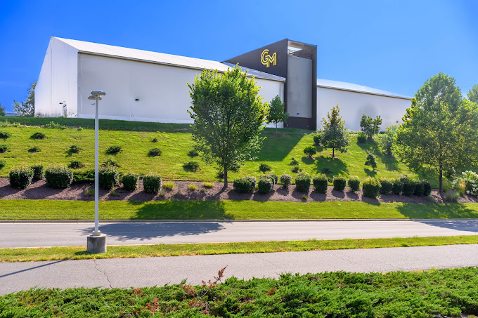 It is a clear day with a blue sky. The new activities building stands in the background at the top of a well cared-for hill with short grass and bushes and trees. It is a large white temporary building with a concrete core structure in the center that stands 40 feet high. The GM logo is prominent on the side of the concrete structure. In the foreground at the bottom of the hill is a road, lamp post, sidewalk, and low lying shrubs.