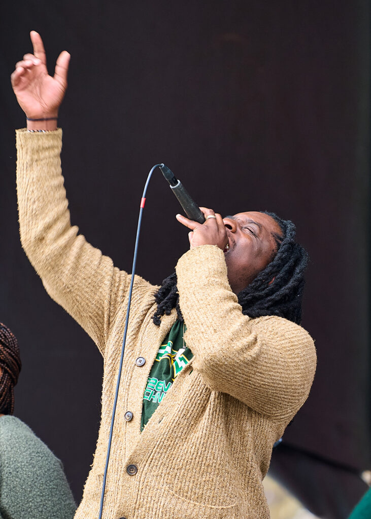 Bobby Lacy raises his right hand in the air as he sings into a microphone in front of a black background. He is wearing a gold button up sweater and a black green machine t shirt.