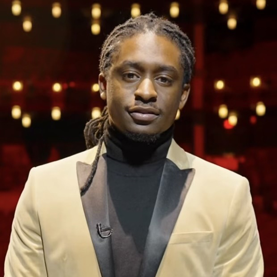 Bobby Lacy is in the Hylton Performing Arts Center on stage with the audience area behind him. The background is red seats ornate columns and warm lights. Bobby is wearing a tan suit and a black turtleneck.