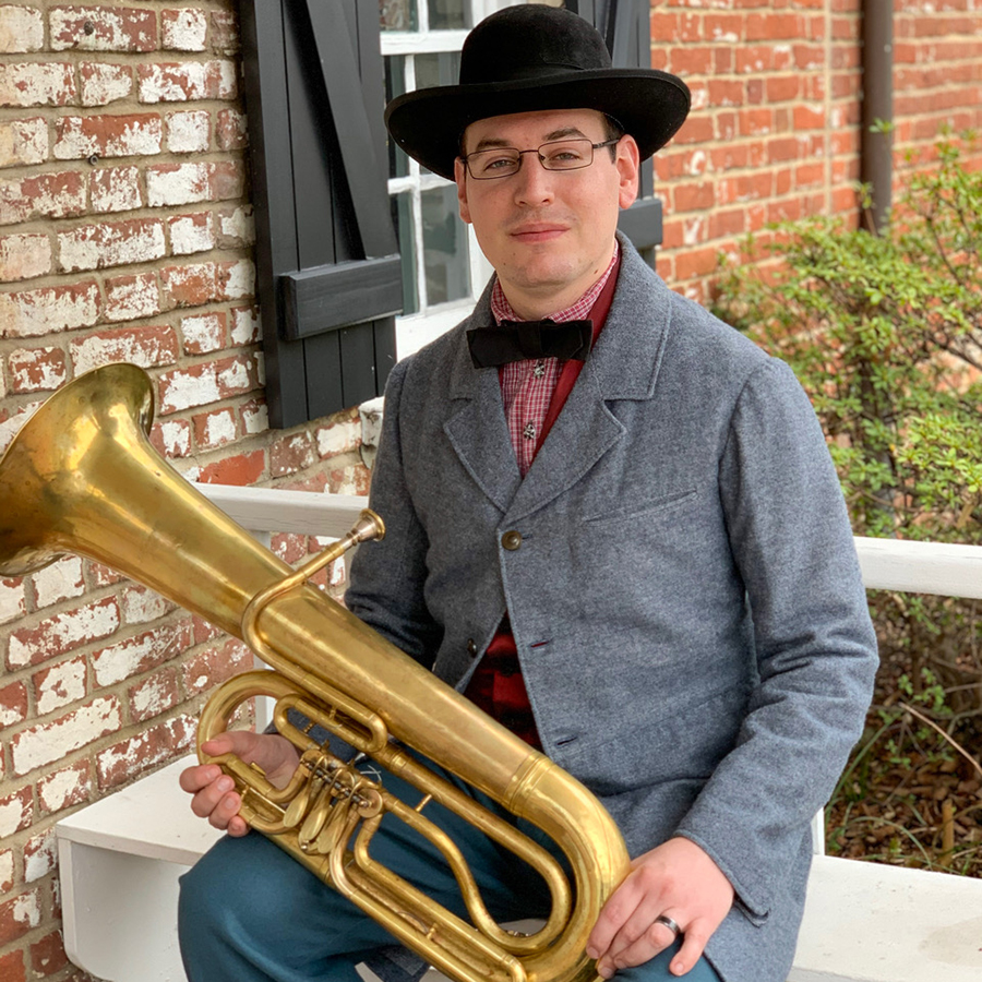 Chris is seated on a white bench on the porch in front of a brick building. The windows have black wooden shutters in a 19th century style. Chris is wearing a 19th century style wide brimmed black hat and gray wool overcoat, a black bow tie and blue wool pants. He is holding a brass keyed euphonium.