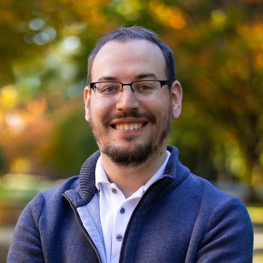 Chris is smiling while standing outside in the fall. The leaves are turning orange and gold behind him in a blurred background. He is wearing glasses, a white button up shirt and a blue zip up sweater.