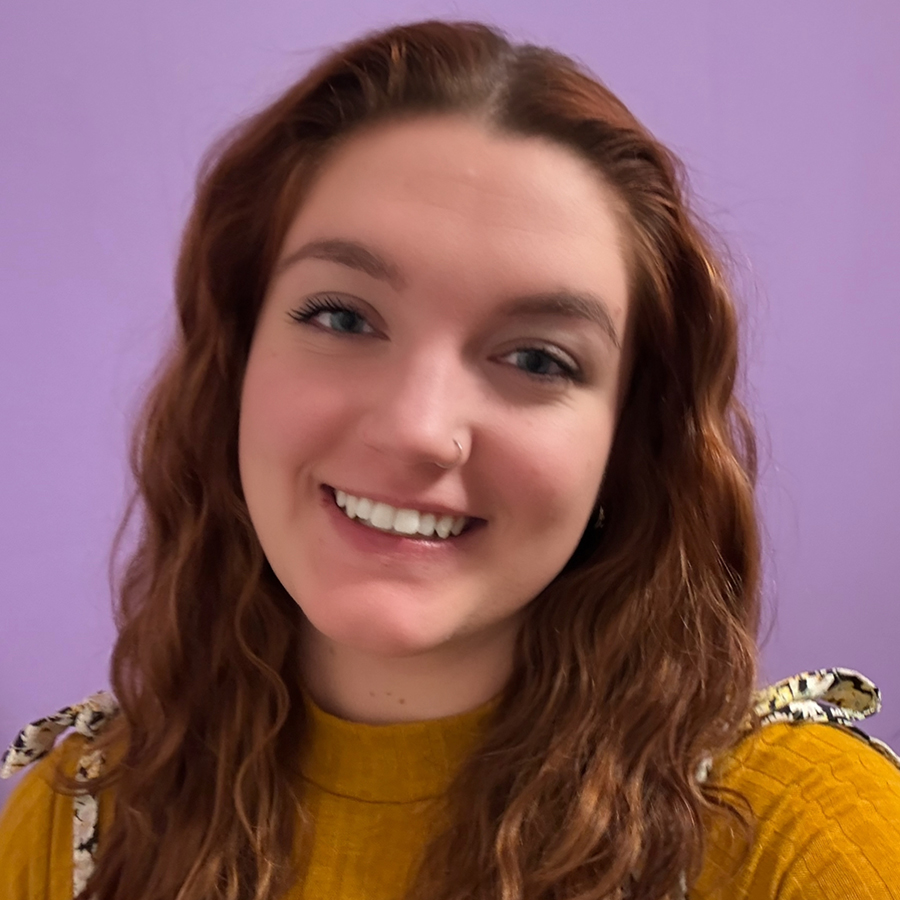 Christine is smiling and sitting in front of a purple background. She has long red hair. She is wearing a mustard colored shirt and patterned overalls.