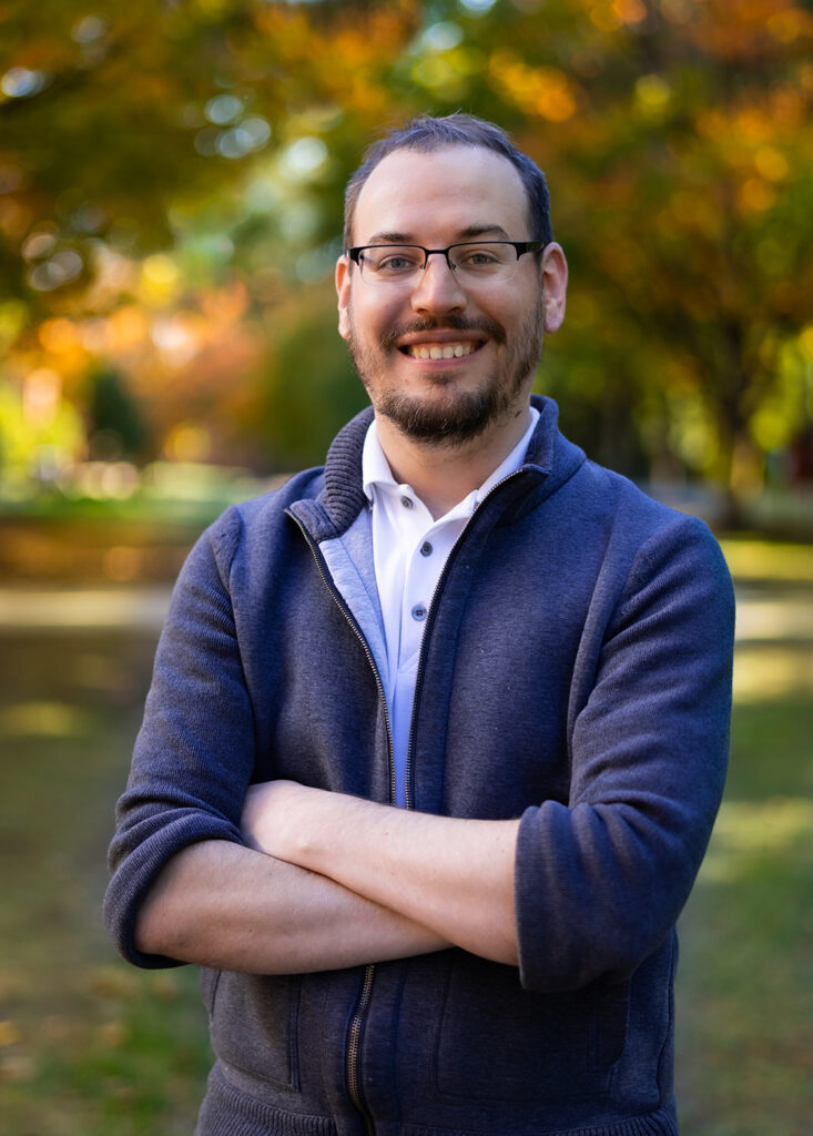 Chris is smiling while standing outside in the fall. The leaves are turning orange and gold behind him in a blurred background. He is wearing glasses, a white button up shirt and a blue zip up sweater.