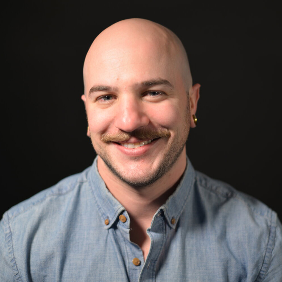 Dan is smiling and sitting in front of a black background. He has a mustache and is wearing a blue collared long sleeve shirt.