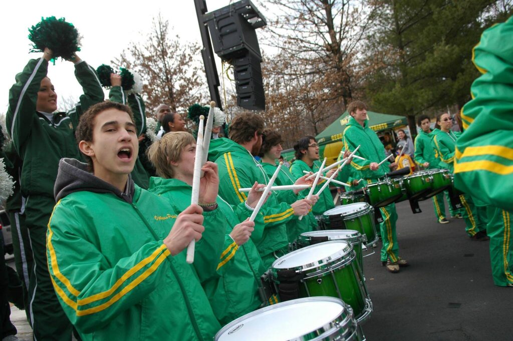 Members of the GMU indoor drumline stand outside during a pep rally near EagleBank Arena. They are all wearing green track suits with yellow stripes running down the sleeves and pant legs. They are chanting and clicking their drum sticks together. Several have snare drums. One member has quad drums.