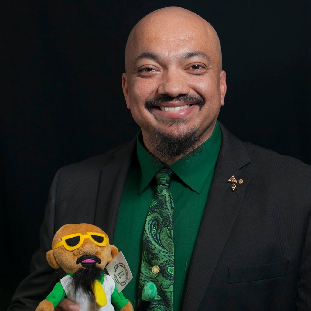 Dr. Nickens is smiling and holding a plushy doll of himself. The background is black. Dr. Nickens is wearing a black suit and a Mason green dress shirt and tie.