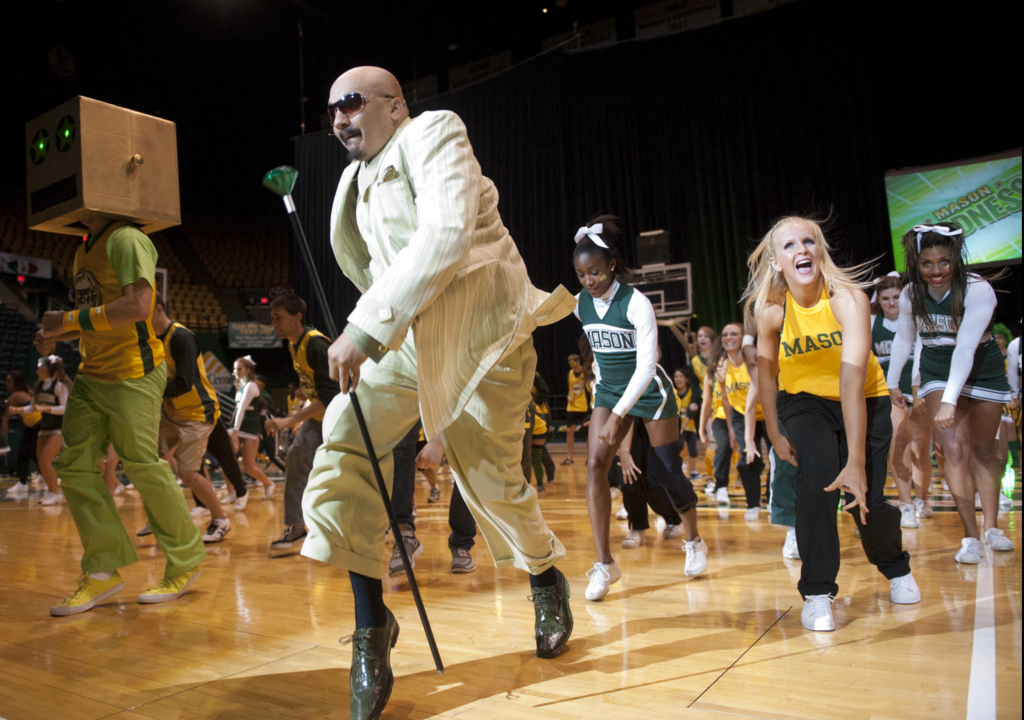Members of the Green Machine, the Mason cheer squad, and the Masonettes dance team are dancing on the basketball court in EagleBank Arena during the Mason Madness pep rally in 2011. Doc Nix is leading the group in a light green and white three-piece suit. Professor Jeremy Freer is wearing yellow chuck's, mint green pants, a yellow basketball jersey and a cardboard box "robot" mask with glowing green eyes. The arena is dark but the floor is lit.