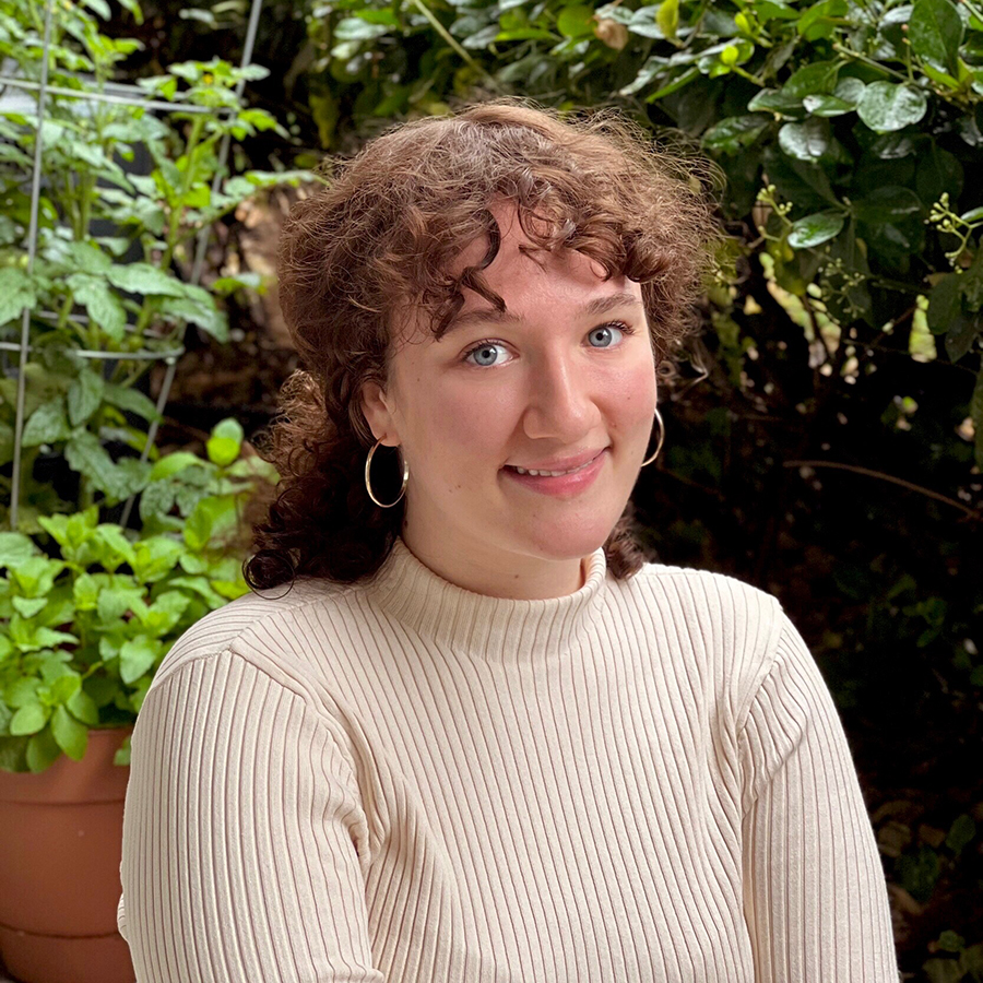 Emma is smiling and sitting on a red seat in front of an array of plants. She has brown hair that is tied back and is wearing a long-sleeve cream colored sweater.
