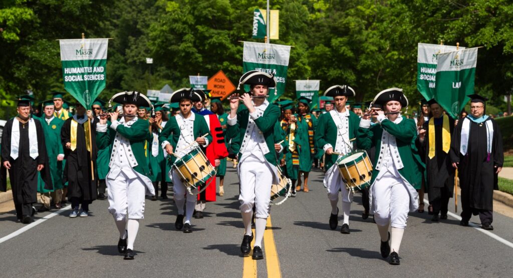 It is morning on the GMU campus. The Patriots Fife and Drum Corps is leading a processional of faculty and students. There are three fife players and three snare drummers mid-performance as they march in formation. They are wearing green and white colonial-era military uniforms with tricorn hats and playing colonial-era fifes and snare drums.