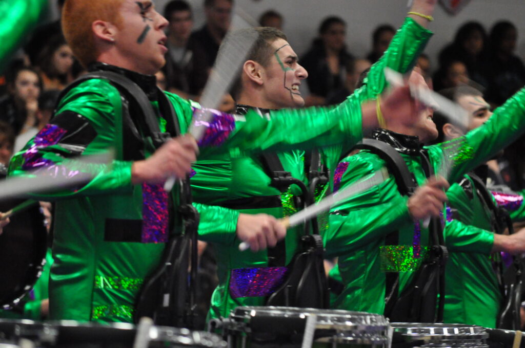 Members of the audience at a competition are in the blurred background. In the foreground, four snare drummers in the GMU Indoor Drumline are mid-performance. Their arms are in sync and moving so fast that they are blurred as they play the drums. They are wearing green sequined uniforms with purple accents and face paint.