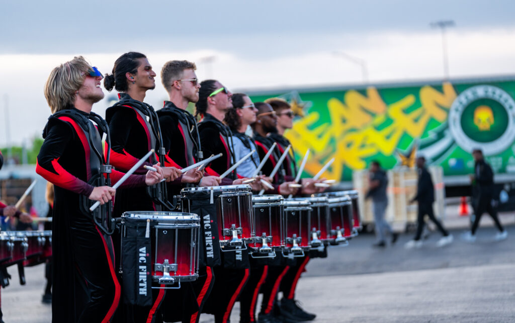 In the blurred background, a box truck is parked. The truck is green and it says "Mean Green" in edgy gold font on the side of the truck. There is also a gold scull and cross-drumstick logo. People are walking in the background. In the foreground, seven members of the GMU indoor drumline are standing in a row. They are wearing black and red stylized uniforms and are playing marching snare drums in unison.