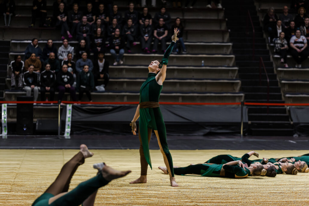 In the center of the floor a guard member reaches to the sky. To their left and right other members lie on the floor as they execute movements. The audience is in the background. The guard is wearing black outfits with green and gold highlights.