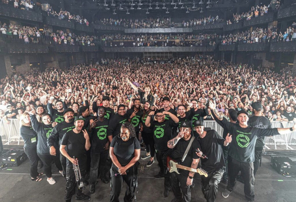 After the show, the anthem audience is in full lights. At the bottom of the image, Griz and members of the Green Machine stand for a group photo smiling on stage. Behind them is a gigantic audience cheering. The band is in black.