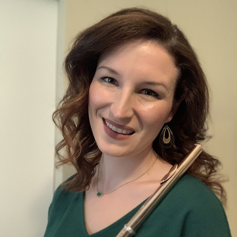 JennaMarie is smiling and standing in front of a cream colored wall. She is holding her flute and has shoulder-length brown hair. She is wearing a green shirt.