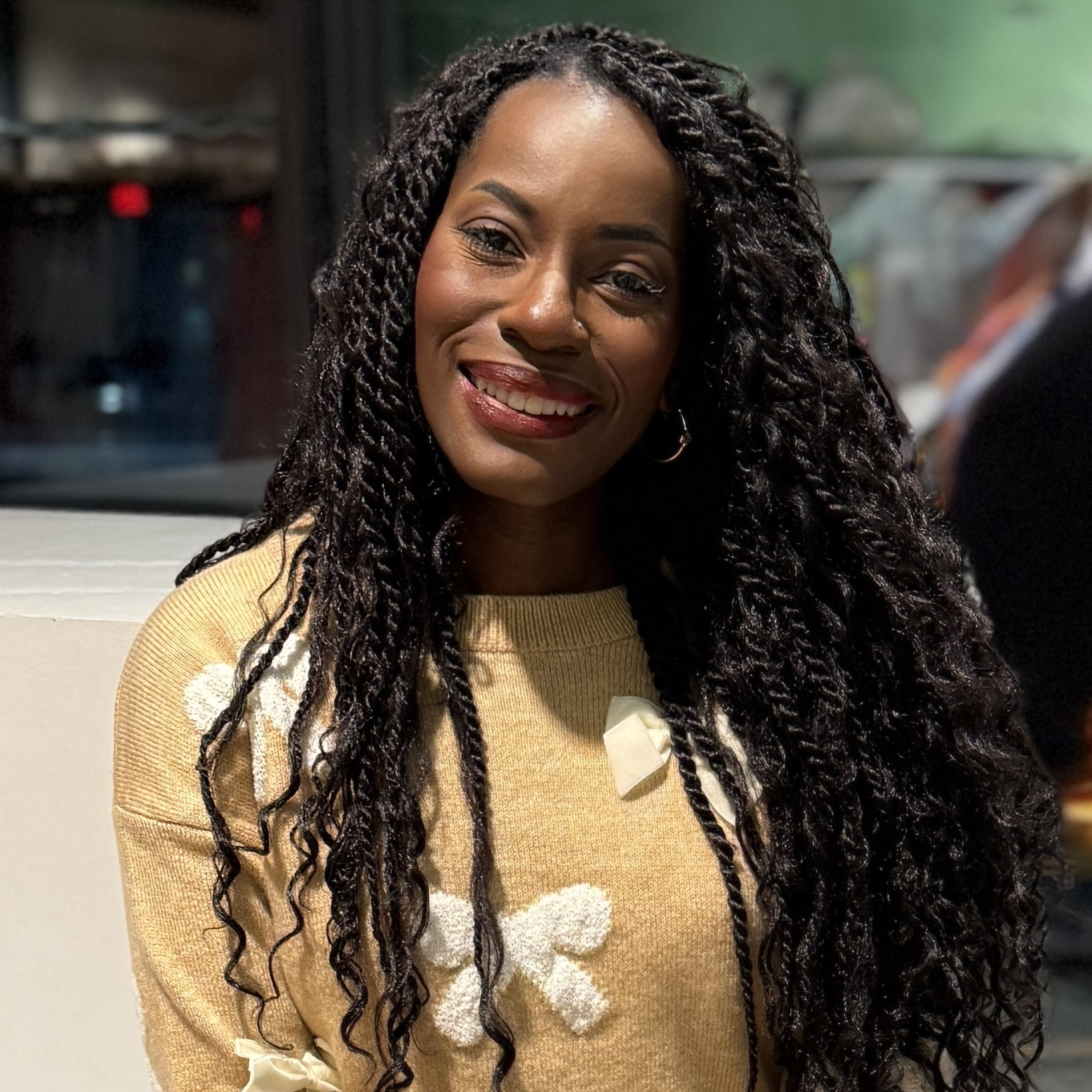 Jennifer is smiling while seated in a blurred theater setting. Jennifer has long black hair. She is wearing a gold sweater with white crocheted bows.