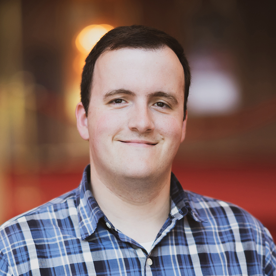 Josh is smiling and seated in a blurred out theater space. Josh is wearing a blue, black, and white flannel shirt.