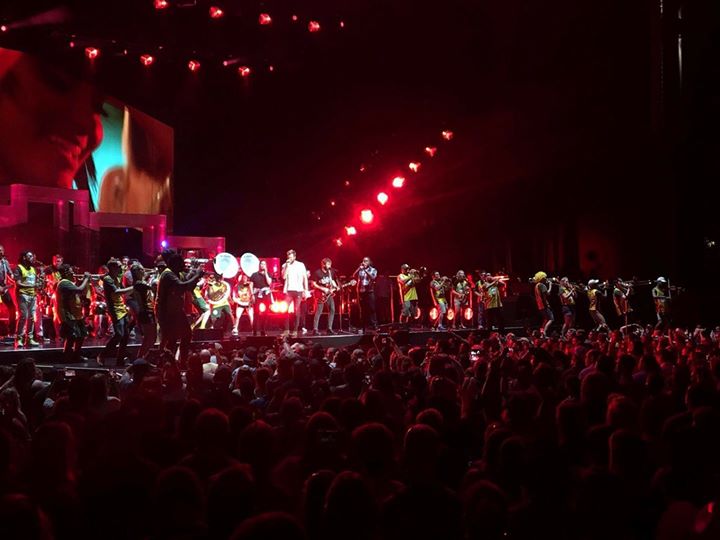 Jiffy Lube live is dark. The stage is lit up with red lights. A gigantic audience is to the right in front of the stage. On stage members of the Green Machine perform with the band Lady A.