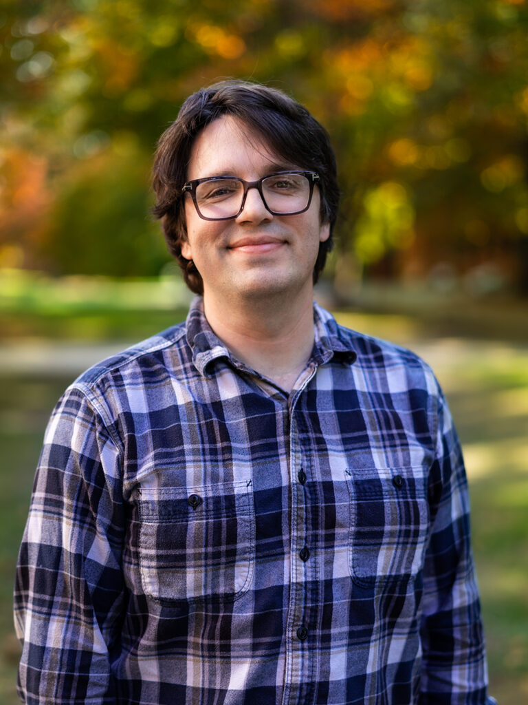 Matt Dievendorf is smiling while outside of the Johnson Center in the Fall. The leaves of the trees behind him are turning orange and yellow. Matt is wearing a blue, gray, and white flannel shirt and glasses.