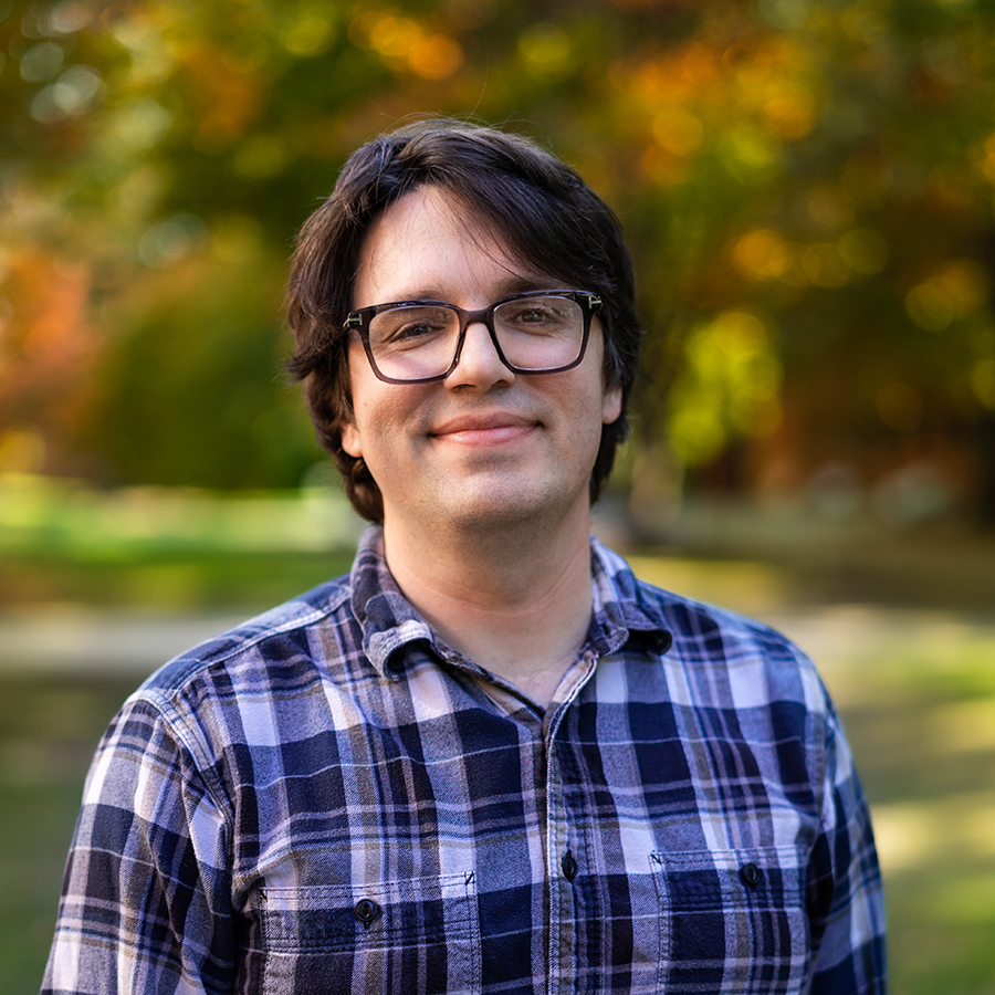 Matt Dievendorf is smiling while outside of the Johnson Center in the Fall. The leaves of the trees behind him are turning orange and yellow. Matt is wearing blue, gray, and white flannel shirt.