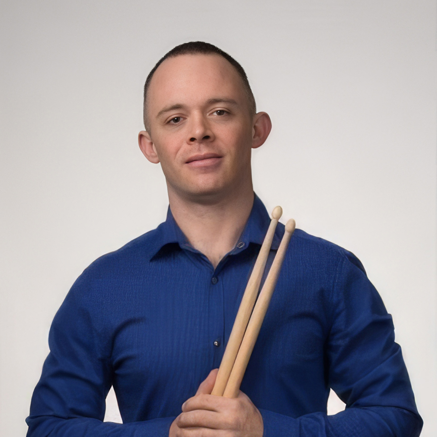 Shane is standing in front of a white background. He has short brown hair, is wearing a fitted blue dress shirt and is holding drumsticks.