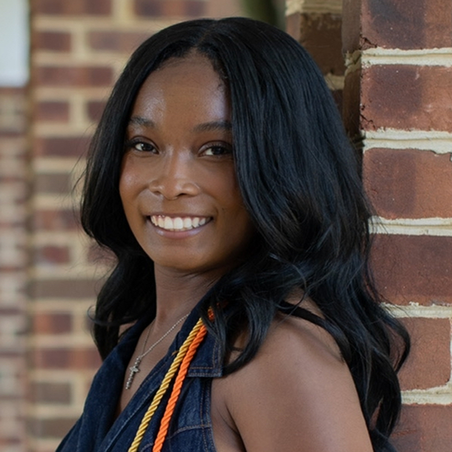 Syla is smiling and standing in front of a brick wall. She has long black hair and is wearing a denim shirt and yellow and orange cords.