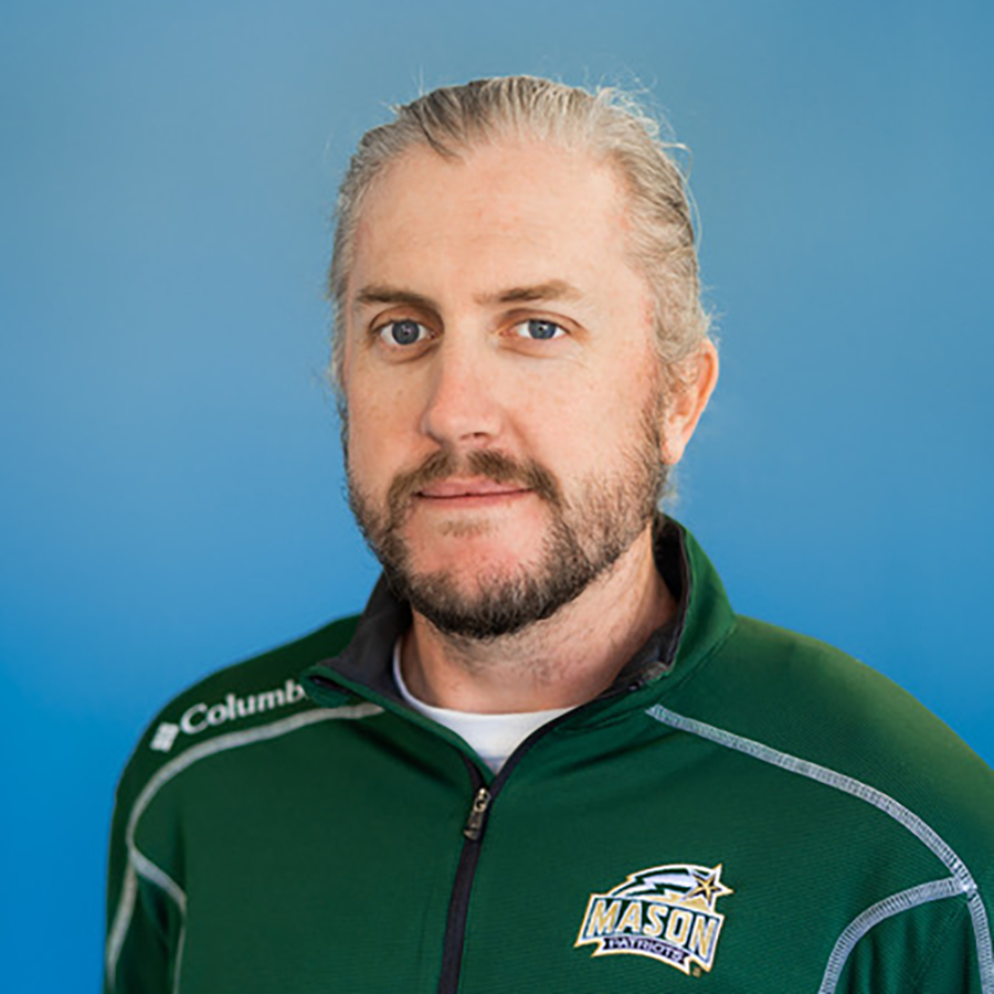 Travis is smiling while seated in front of a blue background. Travis has long blond hair pulled back and a beard. Travis is wearing a green Mason and Columbia branded zip up long sleeve shirt.
