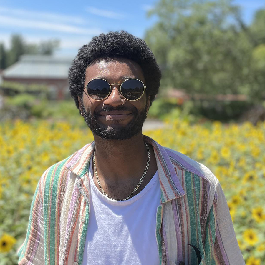 Tyler is smiling and wearing dark sunglasses. He is outside mid-day in front of a field of yellow flowers that is out of focus. He has black hair and is wearing a multi-color striped collared shirt over a white t-shirt.