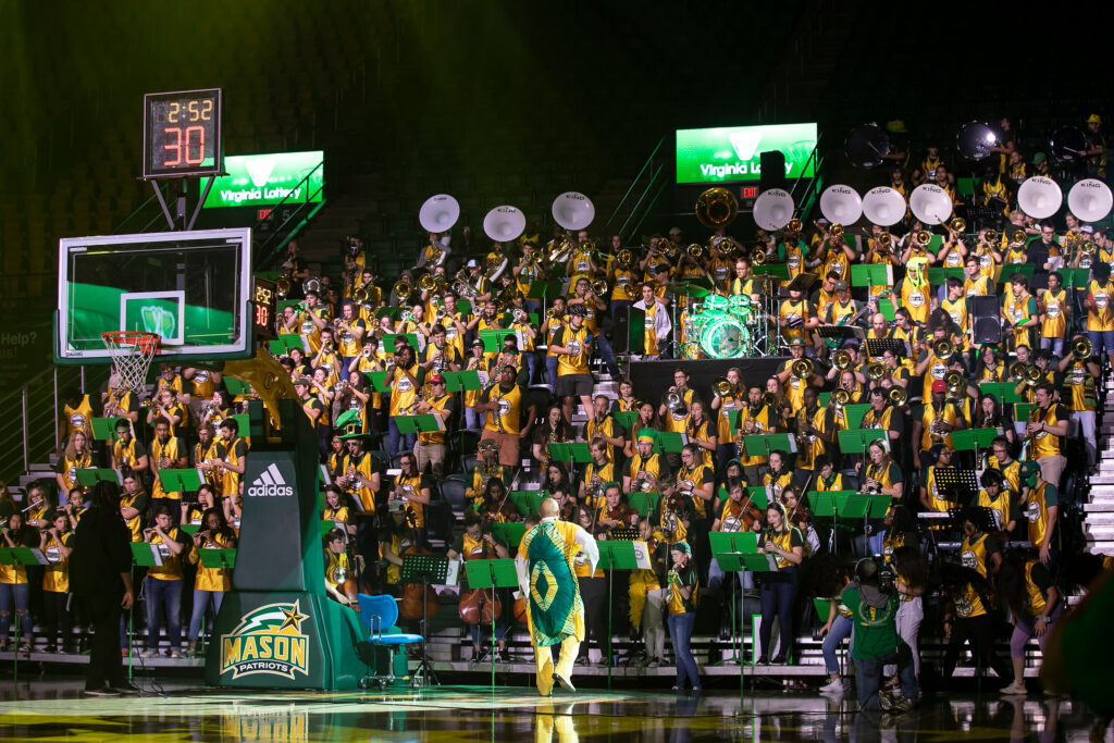 The Green Machine Pep Band performs in the stands in EagleBank Arena during a basketball game. The band is cheering along with cheerleaders in white uniforms. The band is in yellow basketball jerseys. The arena is full of fans screaming.