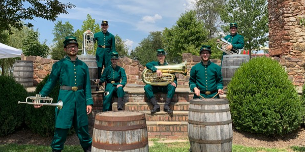 Members of the 8th regiment sit and stand in a candid photo with wooden barrels as props. They have their instruments and are wearing green 19th century military uniforms. They are outside on a sunny day.