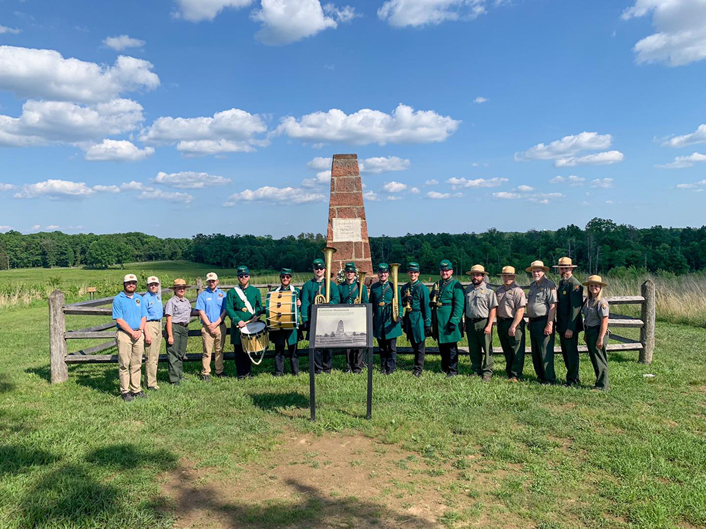 The 8th Regiment band stands in front of a monument at the Manassas battle field on a partly sunny day with park officials for a group photo. The monument and a wooden fence are behind them. They are wearing green 19th century military uniforms and caps and holding their instruments.