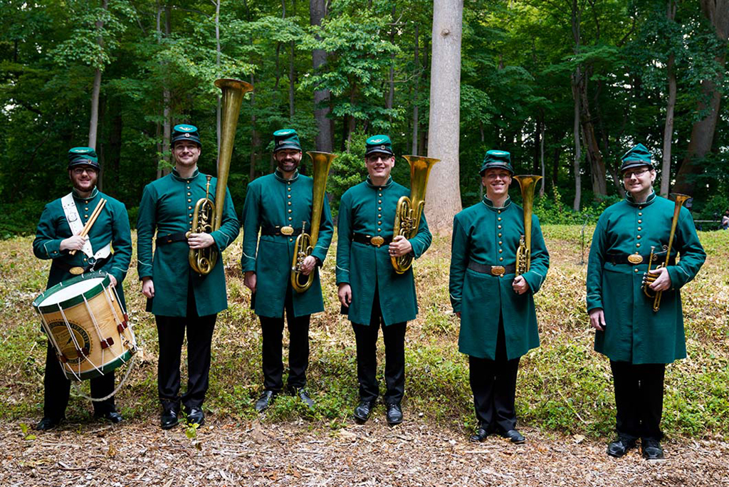 Six members of the 8th regiment band stand at attention in the woods for a group photo with their instruments. They are wearing green 19th century style military uniforms and caps.
