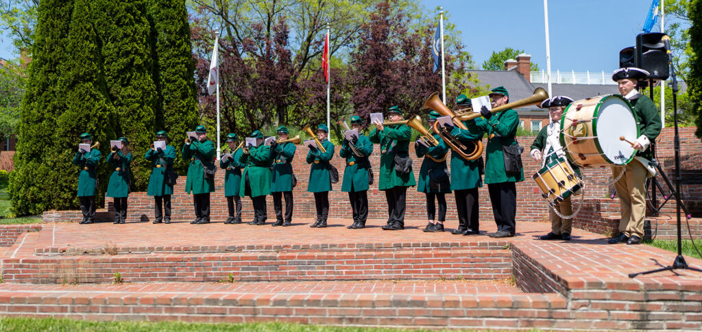 The 8th Regiment band stands in a neat line on a brick stage in an outdoor auditorium. They are playing 19th century horns and drums. It is a bright blue day. The band is wearing green wool coats, green caps, and black pants.
