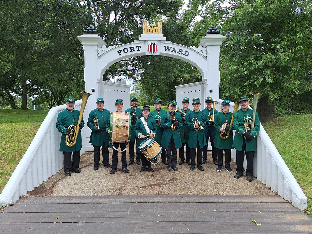 The 8th Regiment band stands in front of the white gate at Fort Ward for a group photo in their green 19th century style military uniforms with green caps and instruments.