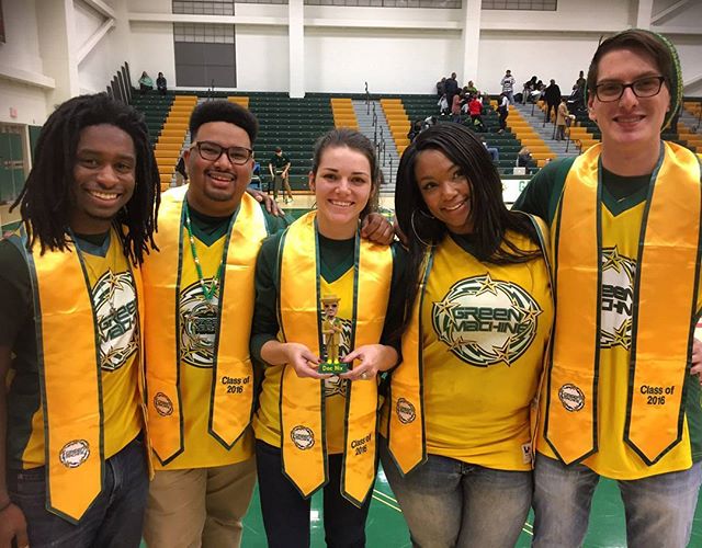 Five green machine alumni stand in an arena for a group photo. They are wearing yellow jerseys and green machine graduation stoles.