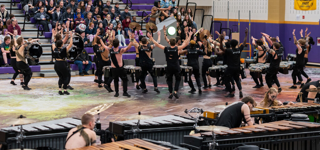 The GMU indoor drumline is mid performance on the floor. They are dressed in torn black post-apocalyptic outfits created for a concept performance and appear to be worshiping a monolith as they perform in front of an audience.