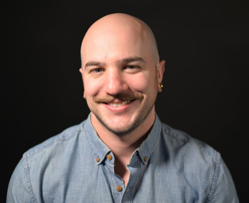 Dan is smiling and sitting in front of a black background. He has a mustache, earrings, and is wearing a blue collared long sleeve shirt.