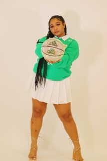 A member of the emeralds stands in a studio for a photo. The background is soft pink white. They are wearing a green cheer outfit with matching print and a white skirt. She is holding a white basketball.