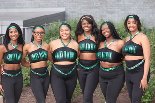 Six members of the Emeralds stand in front of a gray campus building for a group photo. They are wearing black dance uniforms with green print that says "Emeralds." 
