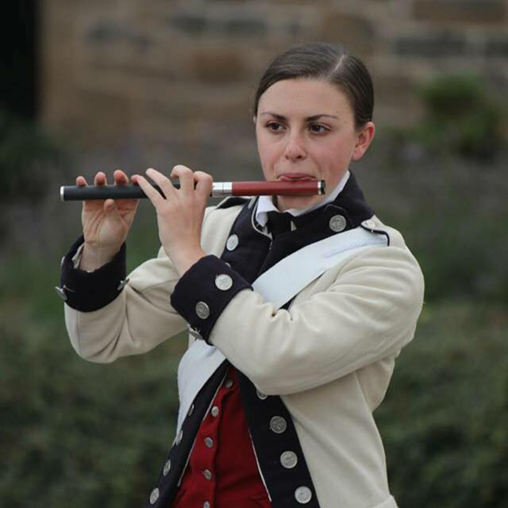 Emily is playing a fife outdoors in front of an out-of-focus brick wall. She is wearing a colonial military uniform that is cream, black, and red. She has a sash from left shoulder to right waist and her hair is pulled back.
