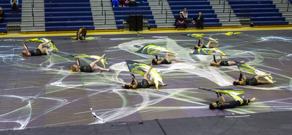 The mason winter guard flag performers lie on the floor as they twirl flags above them. The floor is decorated with a tarp painted with white arcs. The group is in green and gold outfits.
