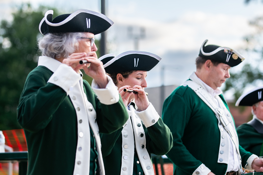 Four members of the fife and drum corps are seen close up in this image outside of the performing arts building at the Fairfax GMU campus. From the waist up they are wearing green and white colonial era military uniforms and black tricorn hats. They are mid performance.