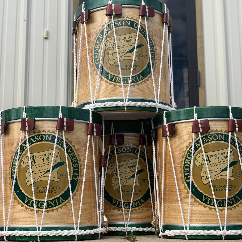 Four colonial era snare drums are stacked in this image in a pyramid formation close up. They are rope drums with green rims and the GMU seal prominently displayed on the sides.