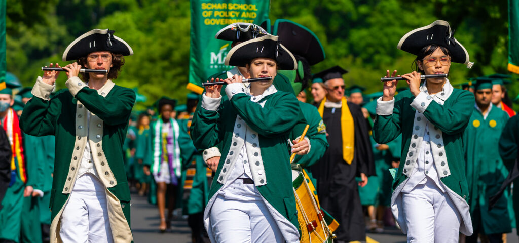 It is morning on the GMU campus. The Patriots Fife and Drum Corps is leading a processional of faculty and students. There are three fife players and one snare drummer mid-performance as they march in formation. They are wearing green and white colonial-era military uniforms with tricorn hats and playing colonial-era fifes and snare drum.