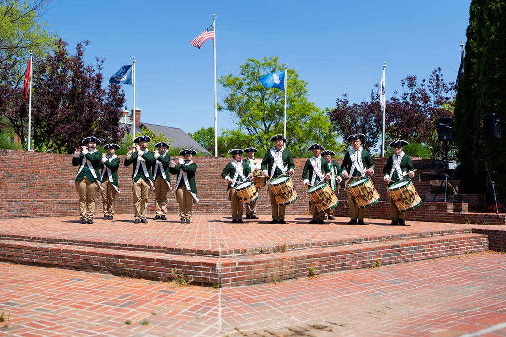 The Fife and Drum corps is standing on a brick stage in an outdoor auditorium on a sunny day. An American flag is on a pole behind them. They are wearing colonial era green and white uniforms and tricorn hats. There are five fife players and seven drummers.