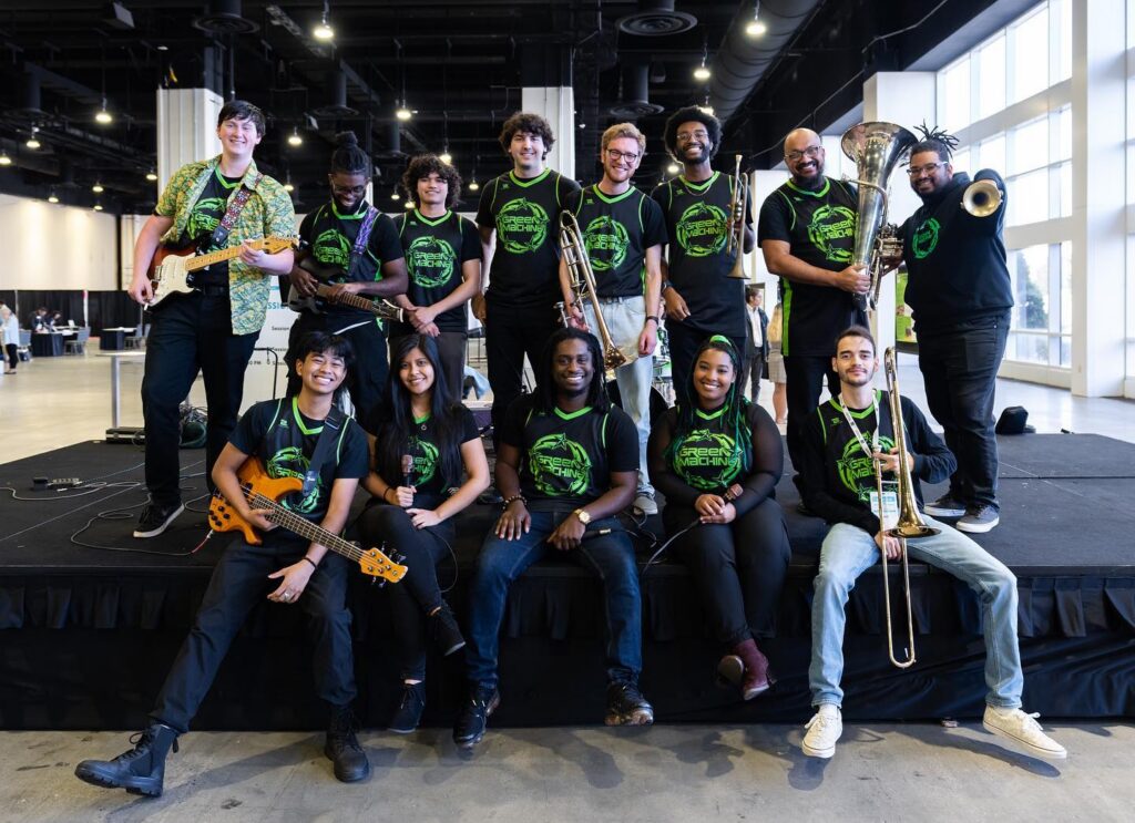 Members of Green and Gold Soul stand and sit for a group photo in an industrial style office space with their instruments. They are wearing black jerseys with green print.