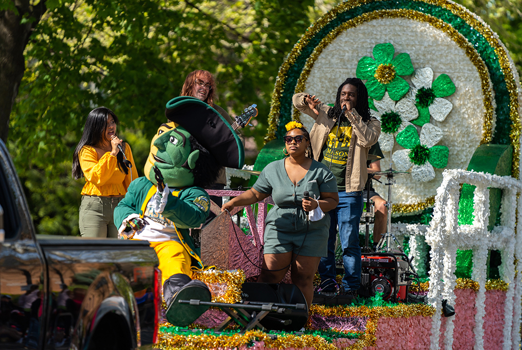 Members of Green and Gold Soul perform on top of an ornate parade float in the Apple Blossom Parade in Winchester, Virginia. The float is being pulled by a black pickup truck. On the float is a drum set player, bass player, vocalists, and the GMUPatriot mascot.