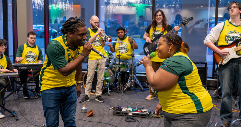 Bobby Lacy and Maya Champion sing into microphones in front of the Green and Gold Soul band inside the lobby of the Center for the Arts on the Fairfax Campus of GMU. The band is wearing yellow jerseys and street clothes. The band consists of a guitarist, pianists, a trumpet player, a bass player, and a drummer.